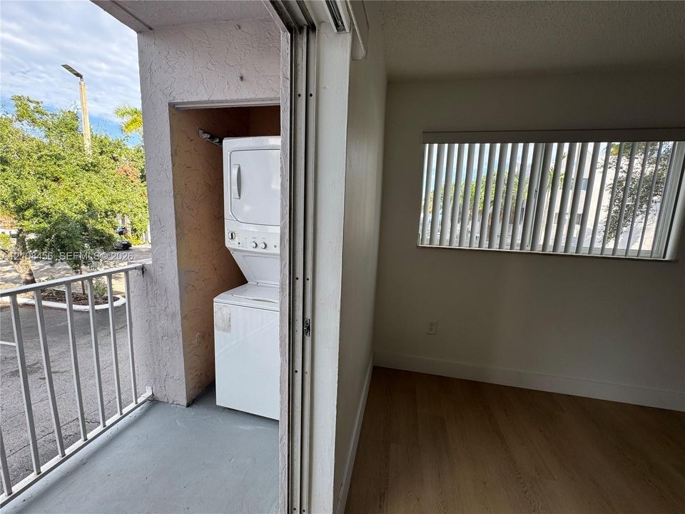 Covered balcony with metal railing, wood-finish flooring, vertical window blinds, and built-in stacked laundry appliance