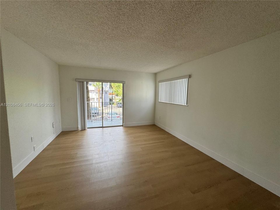 Interior space featuring light wood-finish flooring, neutral wall tones, vertical window blinds, and sliding glass doors with balcony access