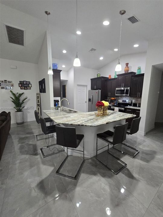 Kitchen featuring dark-finish cabinetry, stone countertops, a multi-seat island with an integrated stainless steel sink, tile flooring, and recessed ceiling lighting