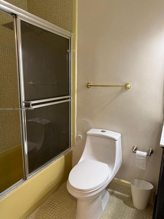 Bathroom featuring a shower-tub combination with sliding glass doors, small-format tile wall cladding, a high-profile white toilet, and gold-finish hardware