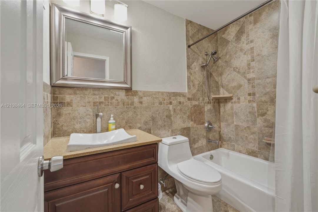 Bathroom featuring a wood-finish vanity with a stone-surface countertop, rectangular vessel sink, wall-mounted mirror, and a tub-shower combination enclosed by tile surround with accent detailing