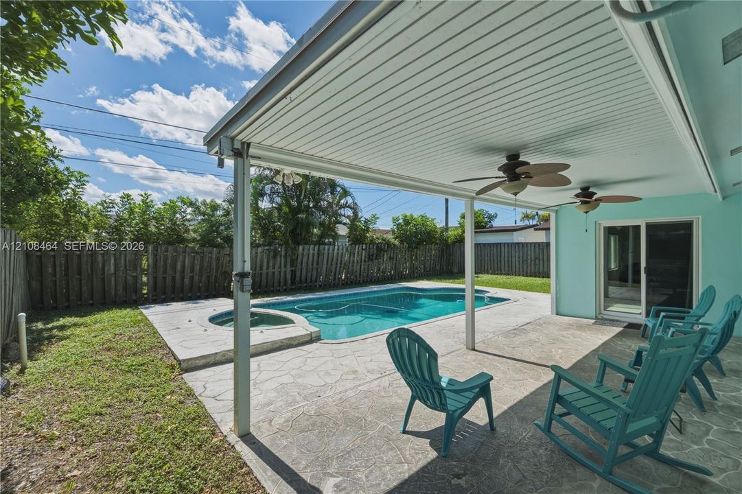 Covered patio featuring stamped concrete flooring and dual ceiling fans, overlooking an inground pool with an integrated spa, all enclosed by a wooden privacy fence