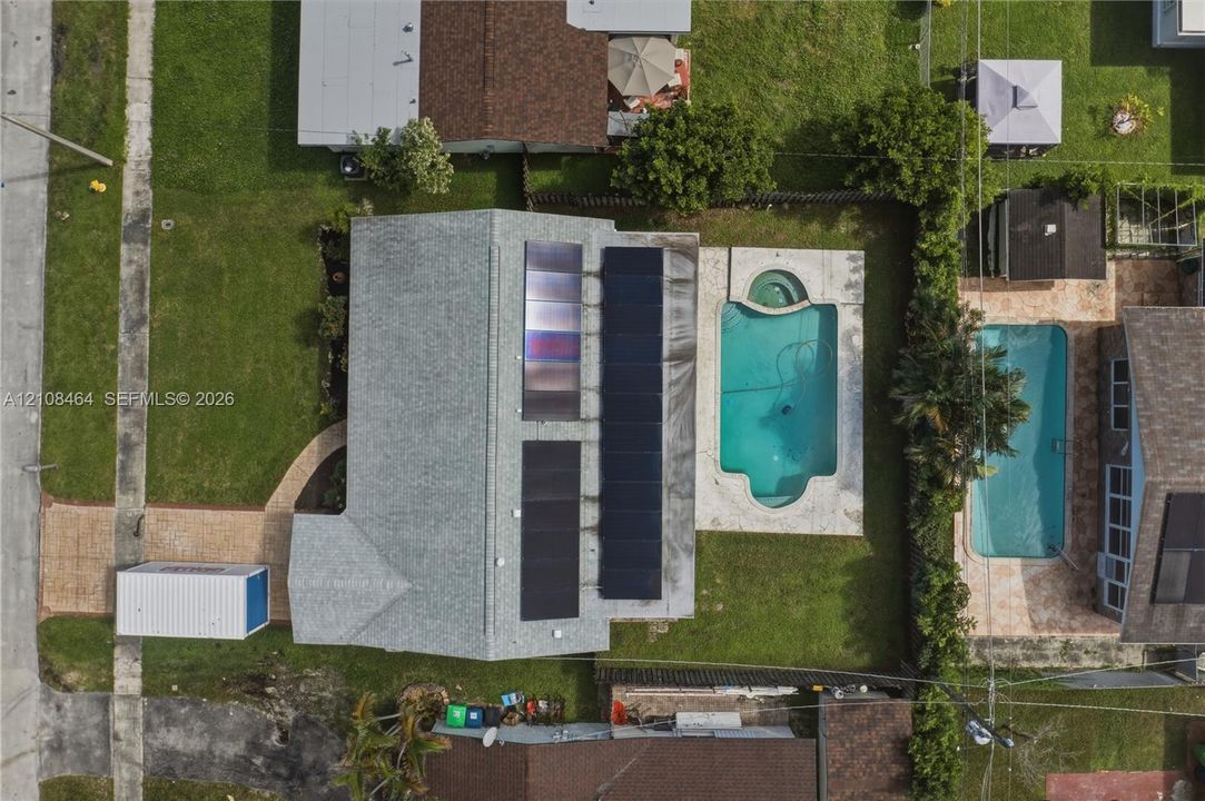 Aerial perspective featuring a shingled roof with solar panel arrays, a rectangular in-ground swimming pool with an integrated spa, and paved walkway