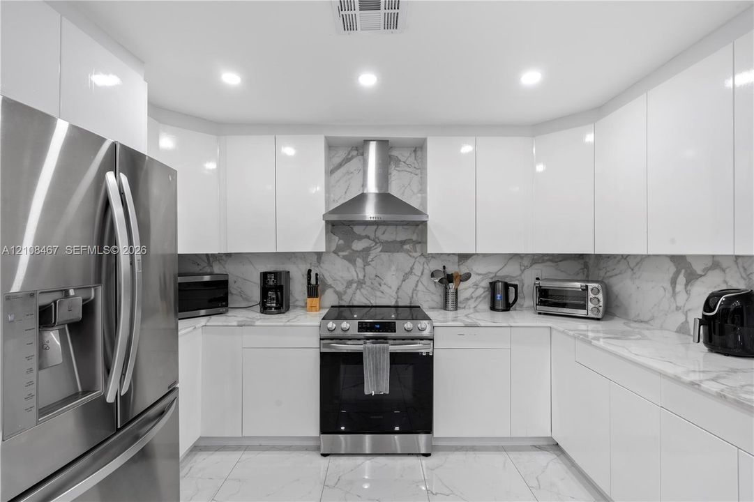 Modern kitchen featuring high-gloss white cabinetry, stone-surface countertops, full-height stone backsplash, stainless steel appliances, and large-format tile flooring