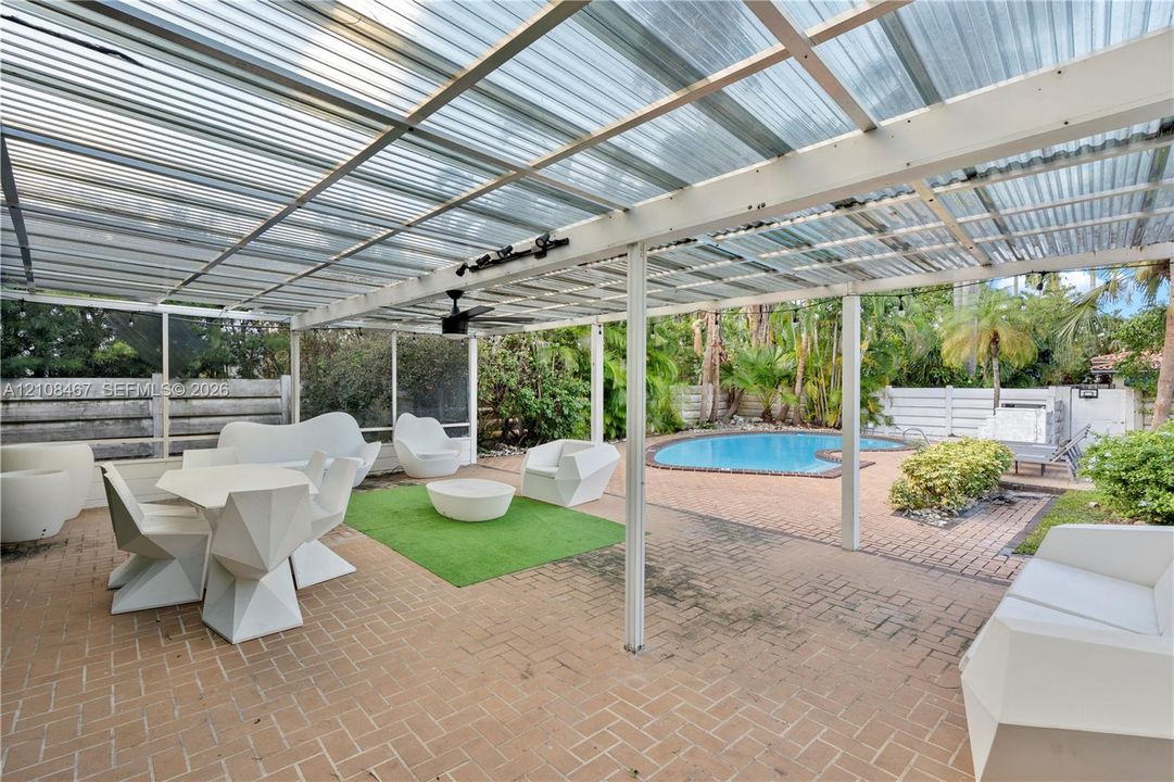 Covered outdoor patio featuring a corrugated translucent roof, integrated ceiling fan, and brick paver flooring, complete with a screened enclosure and an in-ground pool