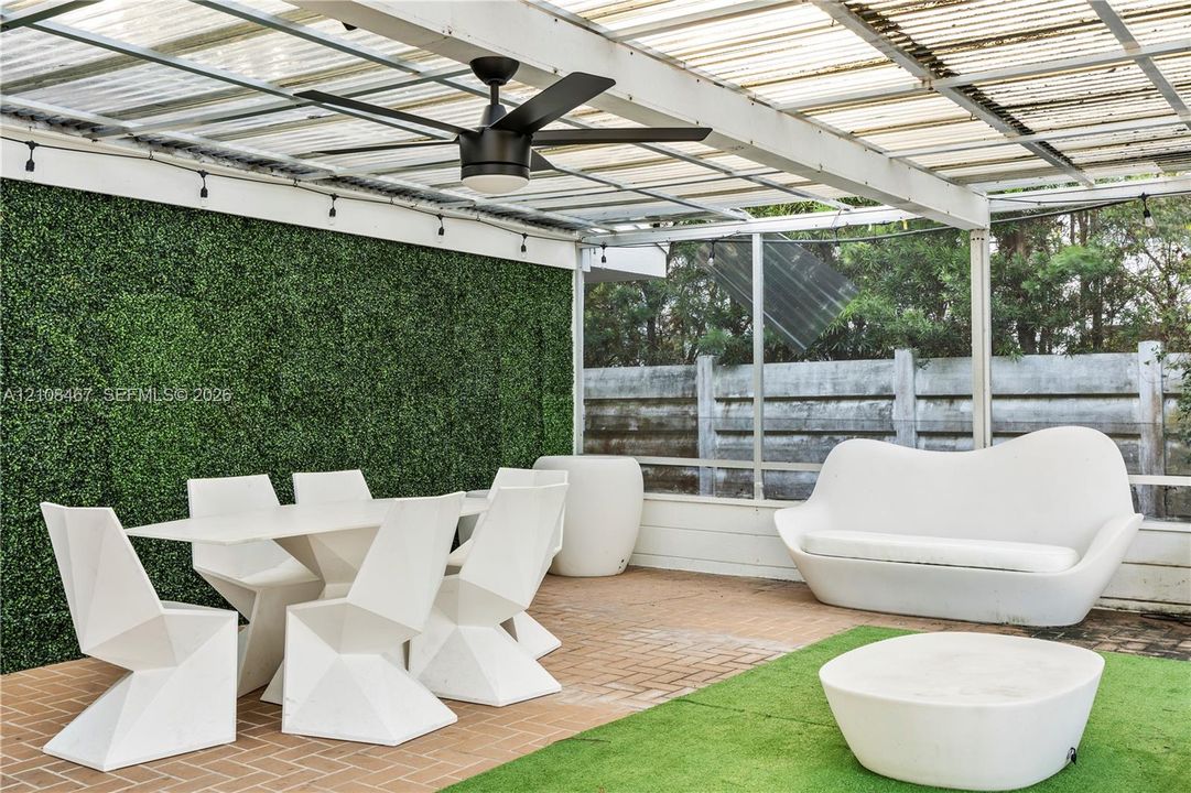 Covered patio featuring a brick-paved floor, artificial grass inlay, a matte black ceiling fan, string lighting, and a green hedge wall