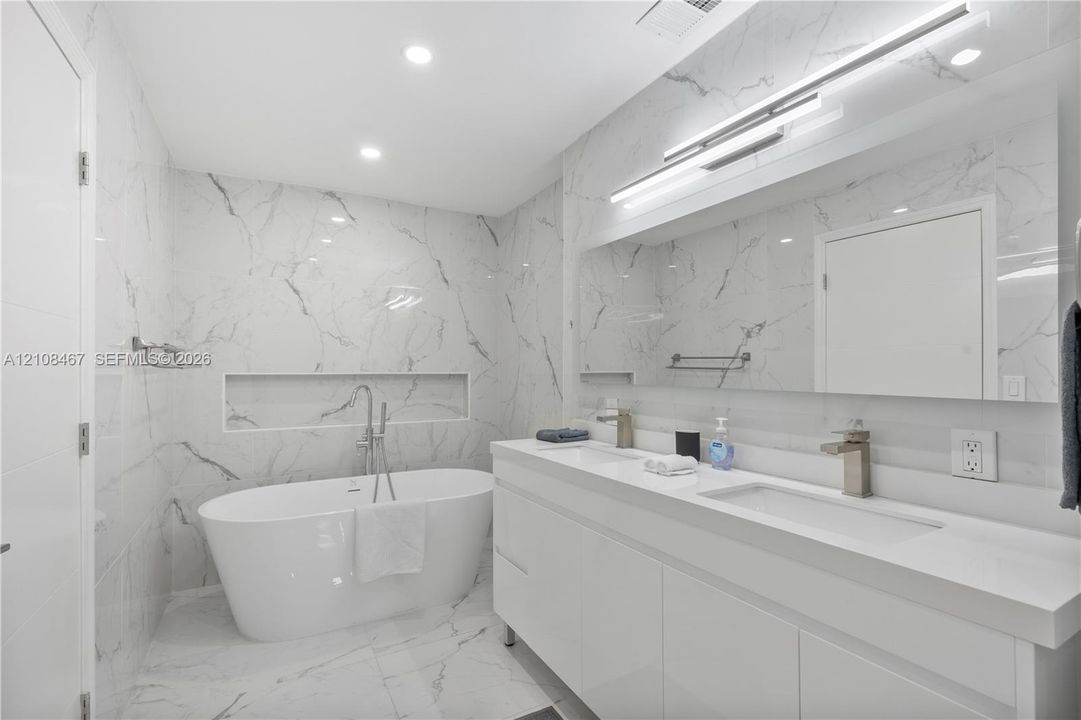 Modern bathroom featuring floor-to-ceiling stone-look tiling, a freestanding soaking tub with a wall-recessed niche, a dual-sink floating vanity, integrated under-cabinet lighting, and a full-width mirror