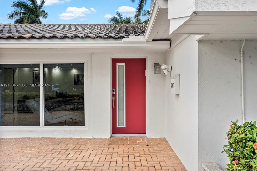 Red entry door with vertical frosted glass insert, terracotta brick patio, white stucco exterior, and barrel tile roof