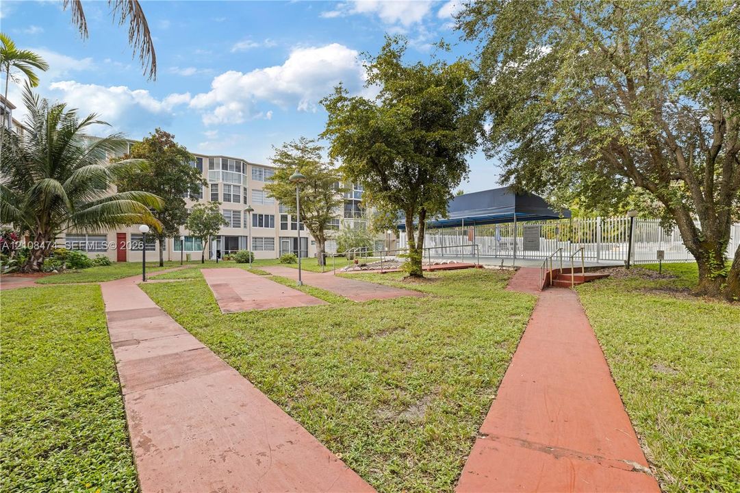 Common area grounds featuring a landscaped lawn, paved walkways, mature trees, and a shaded pool enclosure