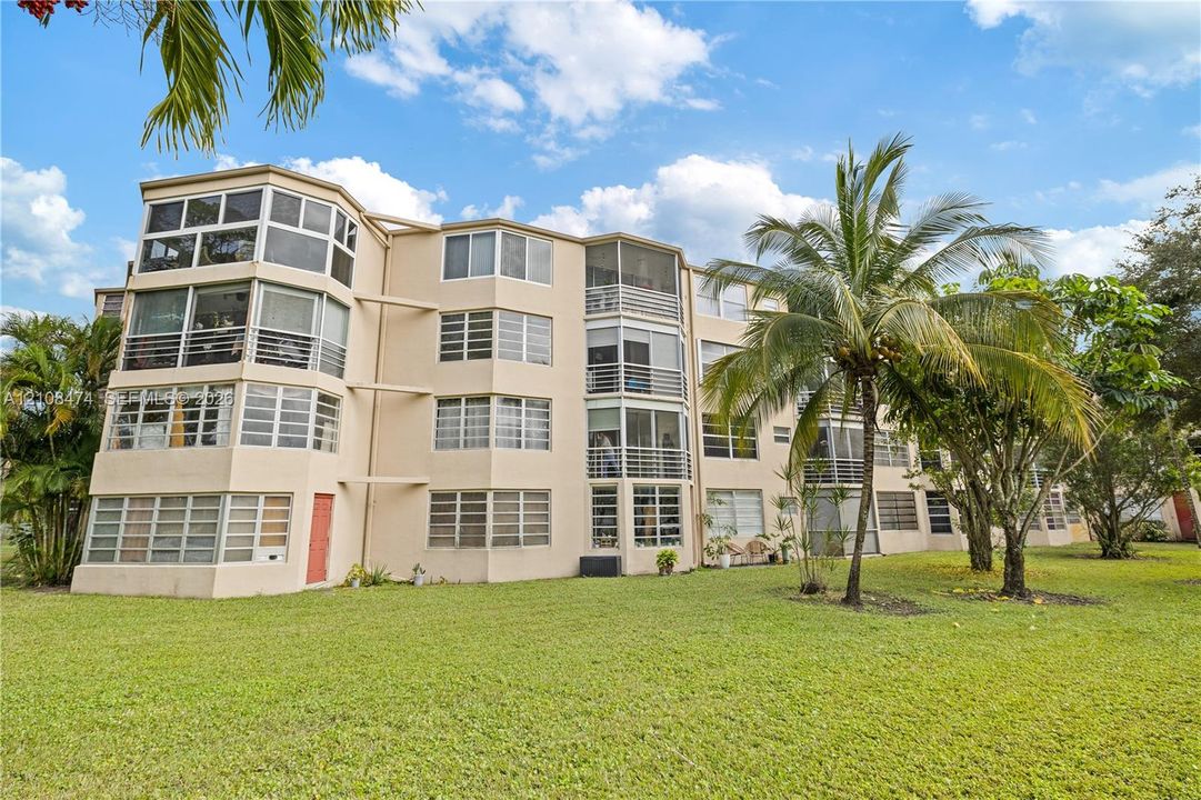 Mid-rise exterior featuring multi-level screened-in lanais, light-toned stucco facade, and expansive lawn landscaping with mature palm trees