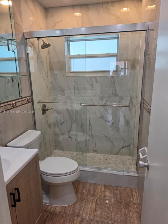 Full-height stone-finish wall tile, frameless glass shower enclosure, integrated pebble-stone shower flooring, wood-finish cabinetry, and polished chrome hardware