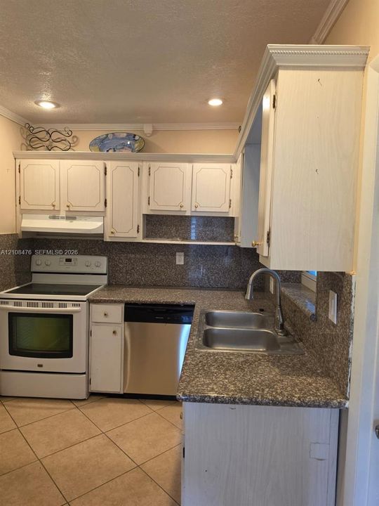 Kitchen featuring white cabinetry with crown molding, stone-patterned countertops, a full-height backsplash, stainless steel dual-basin sink with arched faucet, and ceramic tile flooring