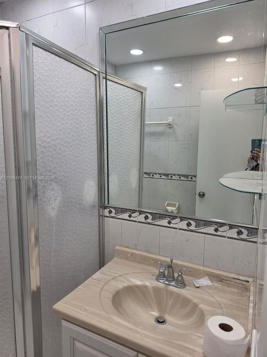 Vanity with an integrated sink and countertop, polished chrome double-handle faucet, oversized wall mirror, walk-in shower with textured glass enclosure, and wall-mounted glass shelving