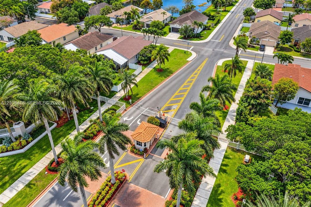 Gated residential entrance featuring a tiled guard house, paved roadways, palm-lined landscaping, and extensive hardscaping