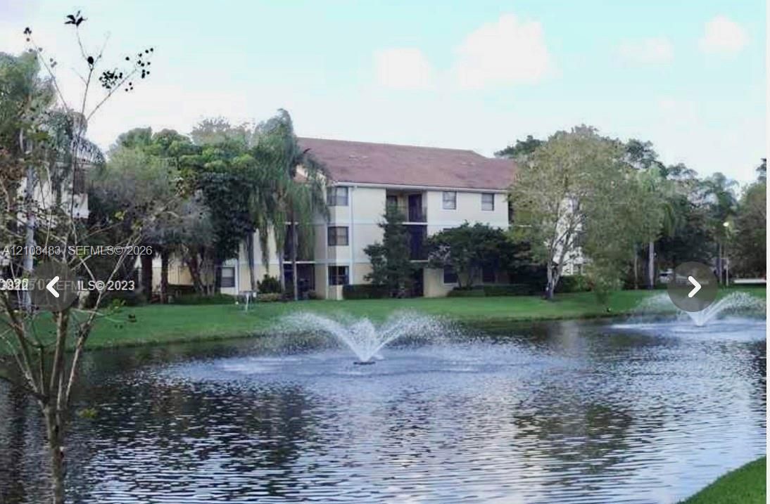 Multi-story building exterior featuring a neutral facade, screened balconies, and a waterfront setting with decorative fountains and established landscaping