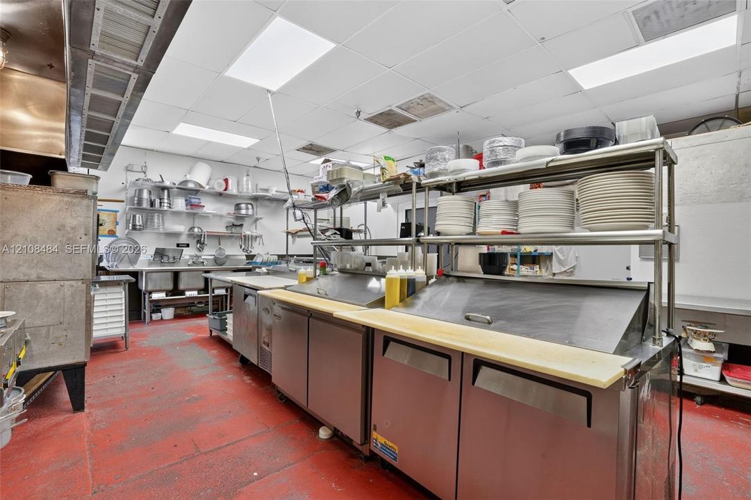 Commercial kitchen featuring stainless steel refrigeration units, multi-tier overhead metal shelving, three-compartment sink, and industrial-grade overhead exhaust hood