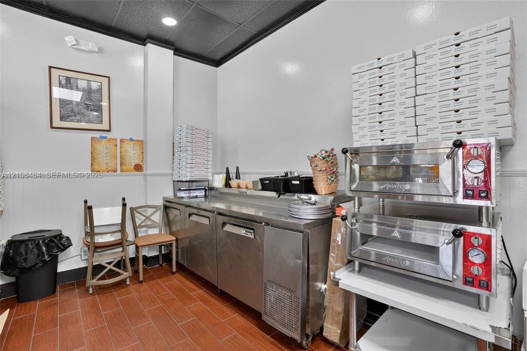 Commercial kitchen space featuring stainless steel prep tables, dual-stack pizza ovens, terracotta-toned tile flooring, and beadboard wainscoting