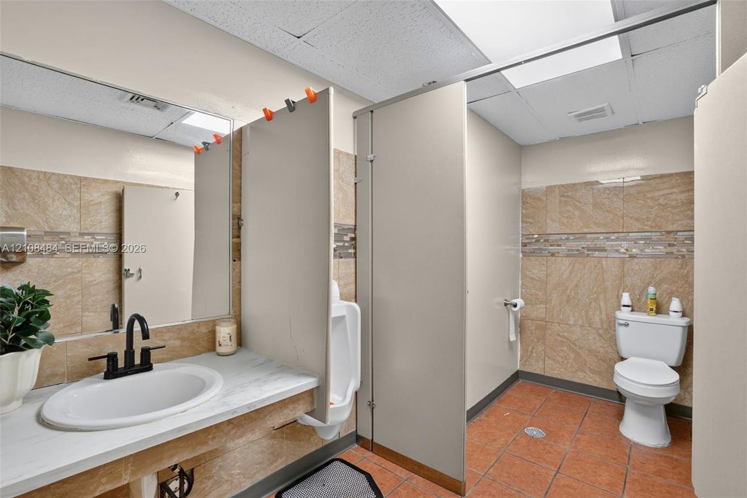 Restroom featuring tile flooring, stone-finish wall tiling with a mosaic accent border, a vanity with a white basin and dark finish faucet, and privacy partitions