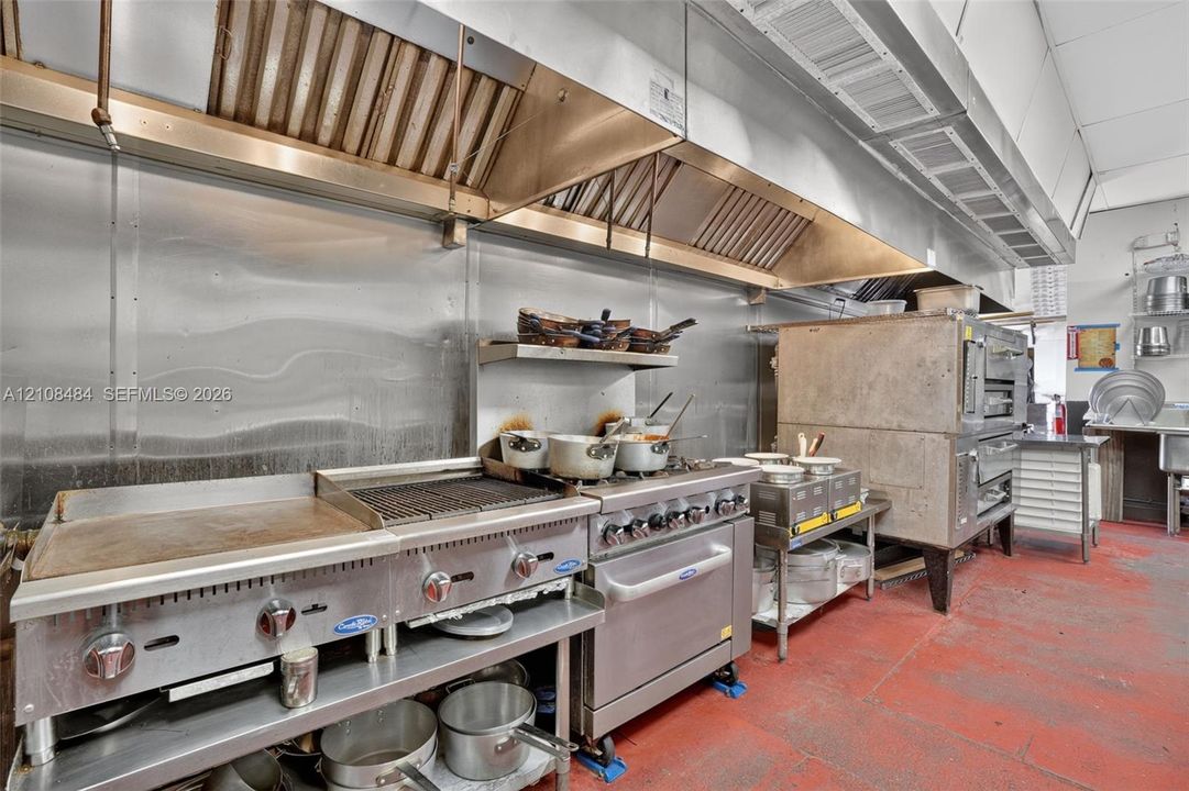 Commercial kitchen featuring a stainless steel flat-top griddle, integrated grill, multi-burner gas range with oven, oversized commercial exhaust hood, and stainless steel backsplash