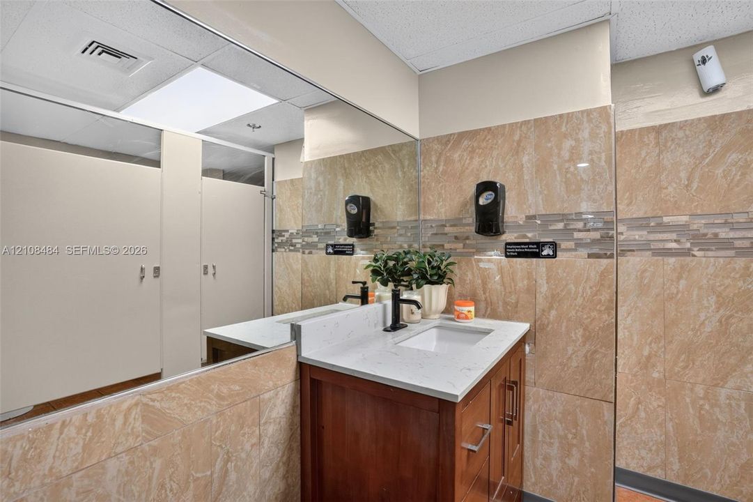 Vanity featuring a light-toned countertop, rectangular undermount sink, and matte black faucet, set against tile-accented walls with an expansive wall-to-wall mirror and privacy partitions