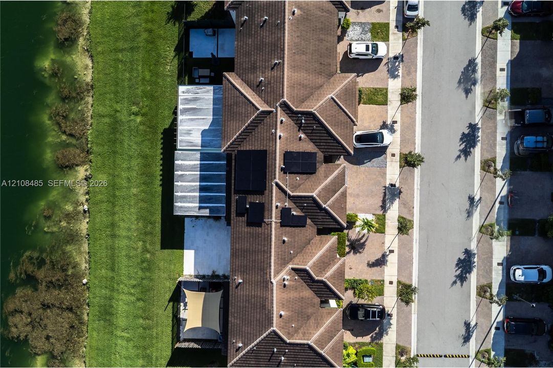 Aerial perspective of a multi-unit property featuring tiled roof lines, integrated solar energy panels, concrete hardscaping, and a grassed area bordering a water feature