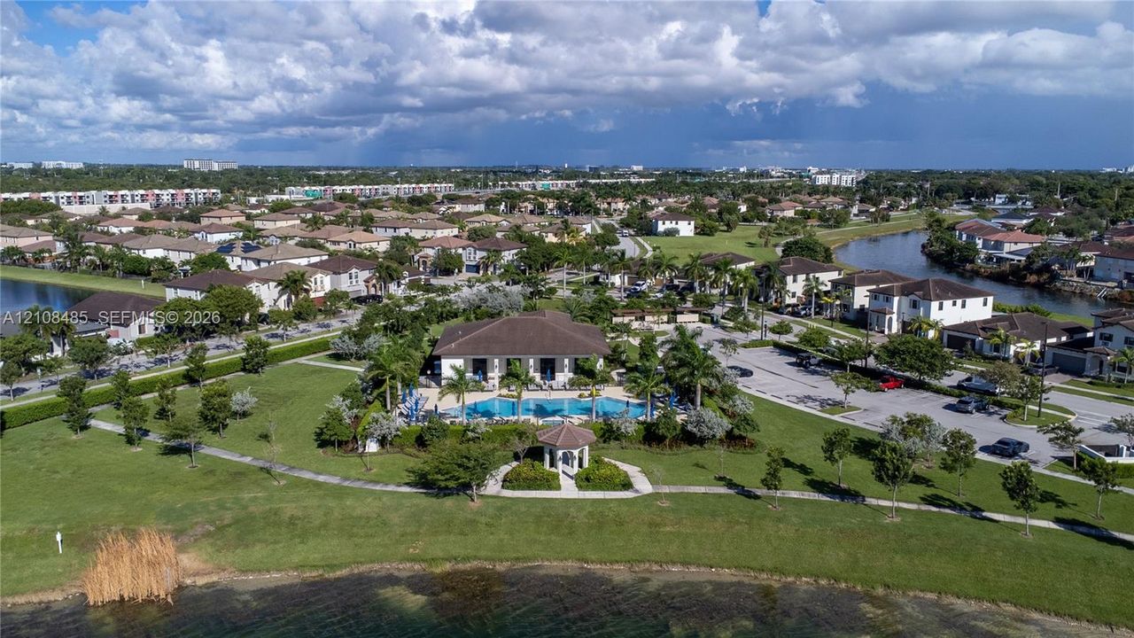 Aerial perspective of a community facility featuring a swimming pool, covered gazebo, surrounding landscaped grounds, and a paved parking area