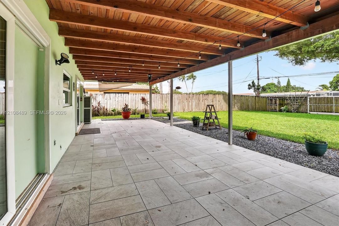 Covered outdoor patio featuring a wood-plank ceiling, large-format tile flooring, and perimeter gravel hardscaping, overlooking a fenced backyard with a lawn