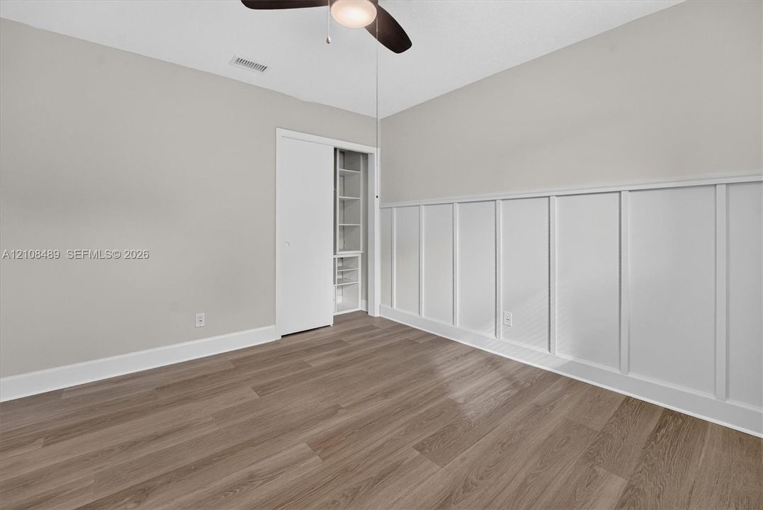 Room featuring white wainscoting, wood-finish flooring, a ceiling-mounted light fixture with a fan, and a closet with built-in shelving