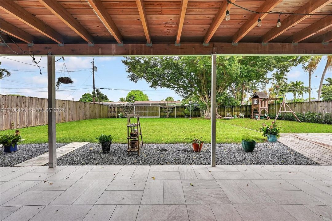 Covered patio featuring an exposed wood-beam ceiling, tile flooring, and a fenced backyard with a lawn, crushed stone border, and mature trees