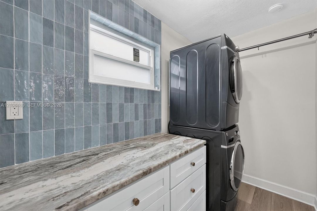 Laundry area featuring white shaker-style cabinetry, stone-patterned countertop, blue vertical subway tile backsplash, and a wall-mounted hanging rod