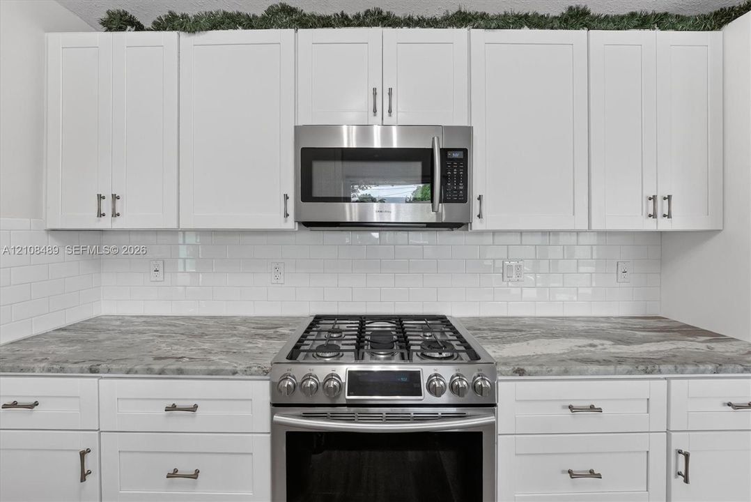 White shaker-style cabinetry with metallic hardware, stone-finish countertops, subway tile backsplash, and stainless steel gas range and built-in microwave