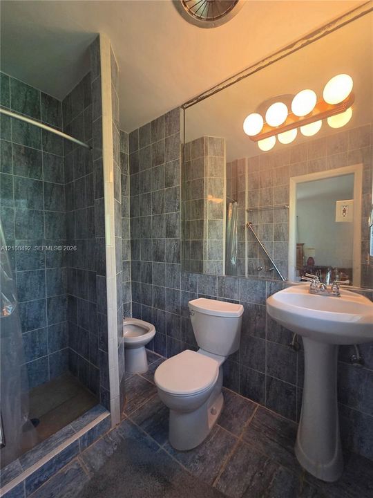 Bathroom featuring floor-to-ceiling stone-finish tile wall surfacing, pedestal sink with double-handle faucet, oversized wall-mounted mirror, and multi-globe vanity lighting
