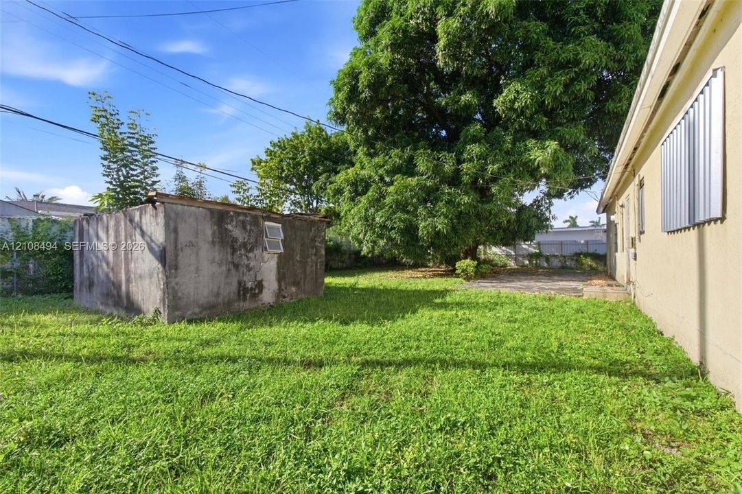 Rear yard featuring a grass lawn, mature tree, masonry outbuilding, and concrete patio area