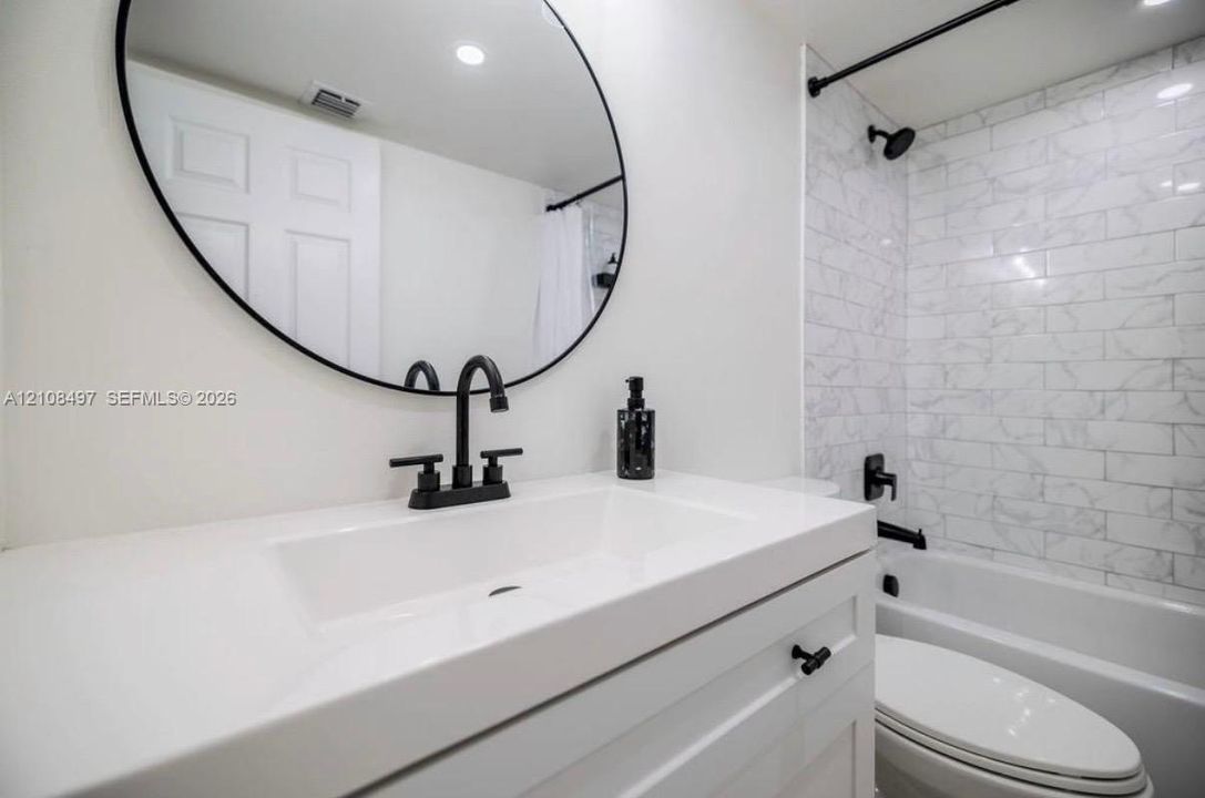 White vanity with a rectangular sink, matte black faucet, oval wall-mounted mirror, and a tub-shower combination featuring subway-tile wall surround
