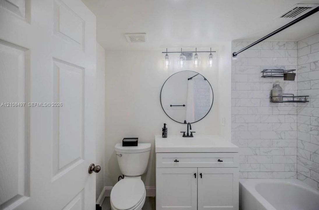 Bathroom featuring a vanity with integrated sink, circular wall-mounted mirror, matte black hardware, and tub surround with subway tile finish