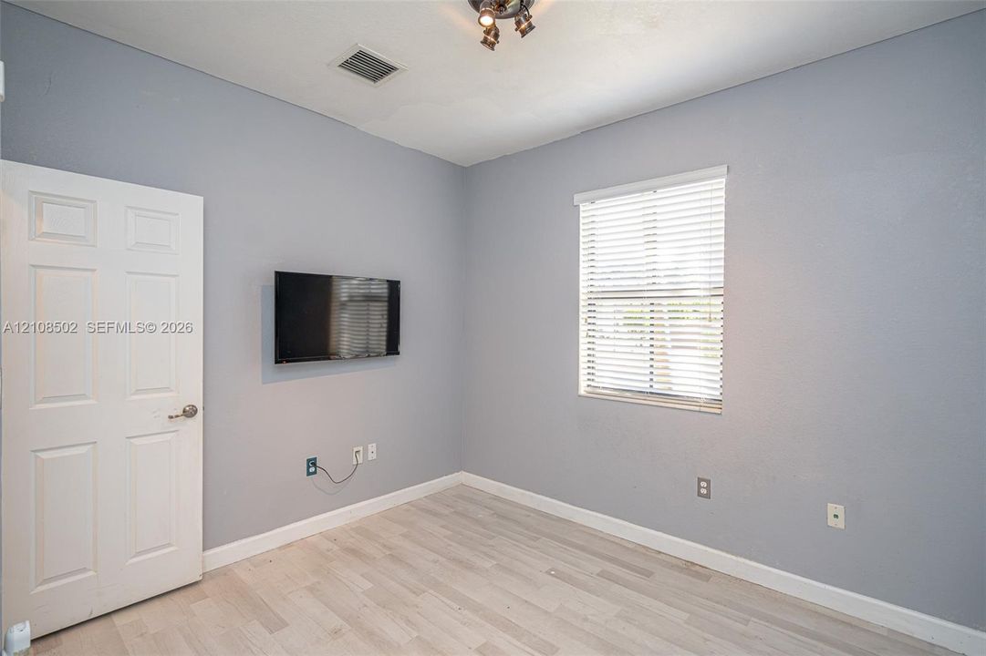 Interior room featuring wood-finish flooring, a multi-panel door with a metallic handle, a window equipped with horizontal blinds, and a mounted adjustable television bracket