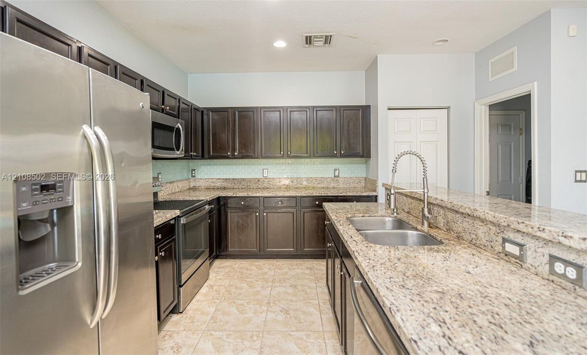 Kitchen featuring dark wood cabinetry, stone-finish countertops, stainless steel appliances, tiled backsplash, and a double-basin undermount sink with a pull-down faucet