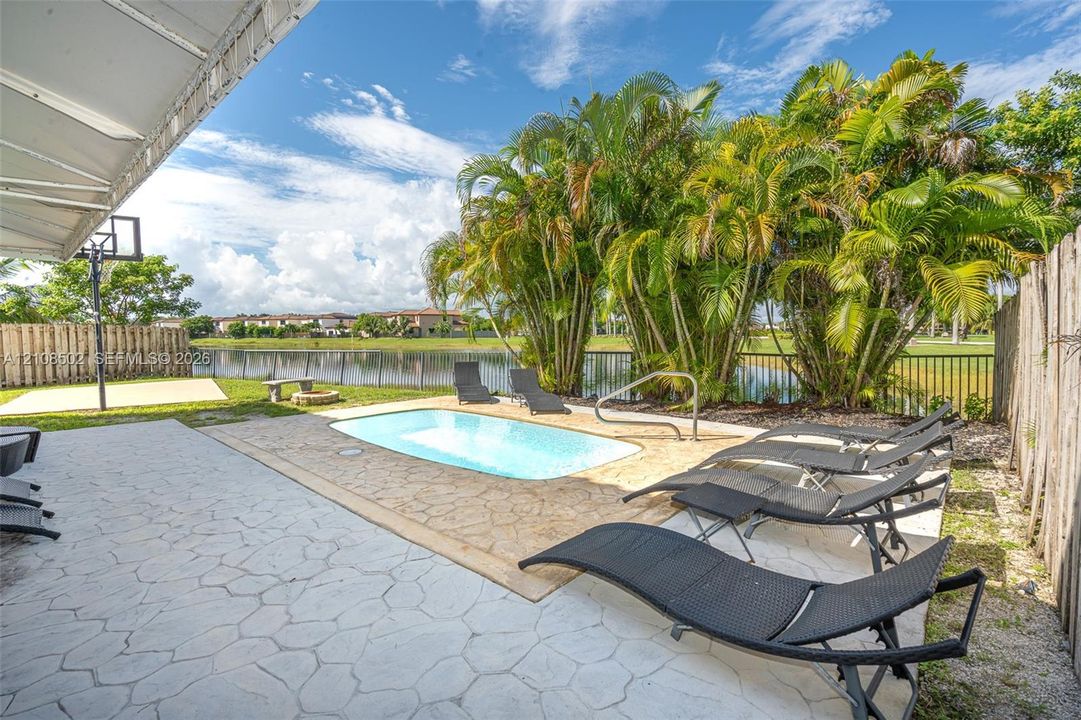 Outdoor living area featuring a paved patio, swimming pool with metal handrail, basketball hoop, and lakefront backdrop