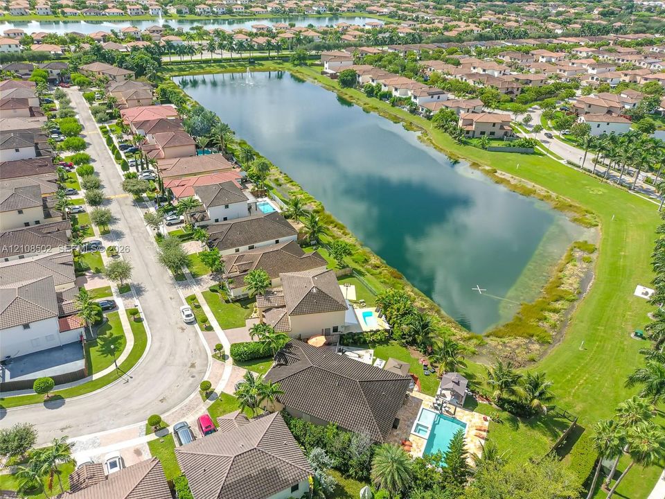 Aerial perspective of a residential property featuring an inground swimming pool, stone-paver pool deck, mature landscaping, and placement along a waterfront pond