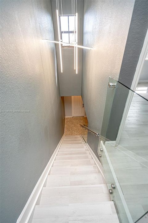 Staircase featuring wood-finish treads, glass panel railing with metallic hardware, and linear LED pendant lighting
