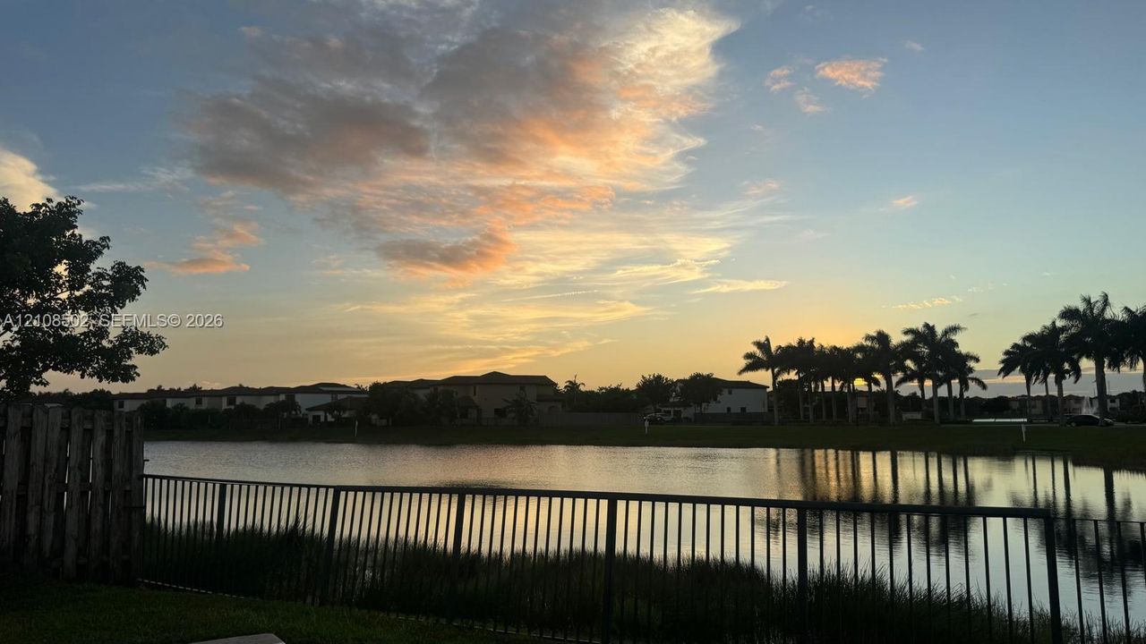 Waterfront property featuring a metal perimeter fence, grassy shoreline, and palm tree canopy