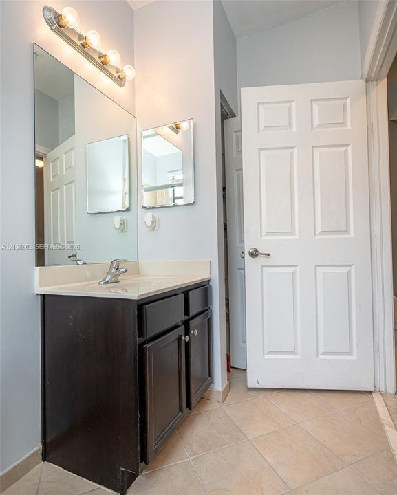 Dark-toned cabinetry vanity with integrated countertop, single-handle faucet, large wall-mounted mirror, four-bulb light fixture, and tile flooring