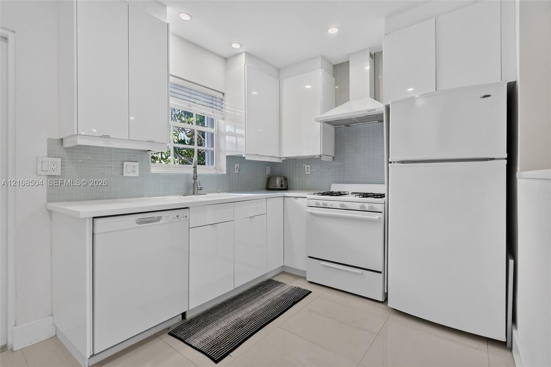 Kitchen featuring flat-panel white cabinetry, light-toned tile flooring, light blue glass tile backsplash, white countertops, and a suite of white appliances