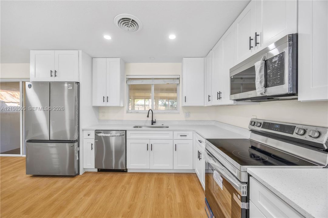 Kitchen featuring white shaker-style cabinetry, solid-surface countertops, stainless steel appliances, matte black hardware, and wood-finish flooring