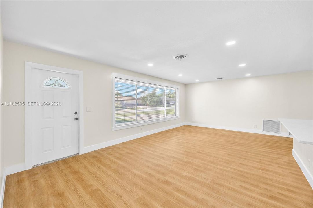 Open-concept area featuring wood-finish flooring, recessed lighting, a wide window with horizontal blinds, and a white entry door with a decorative glass insert
