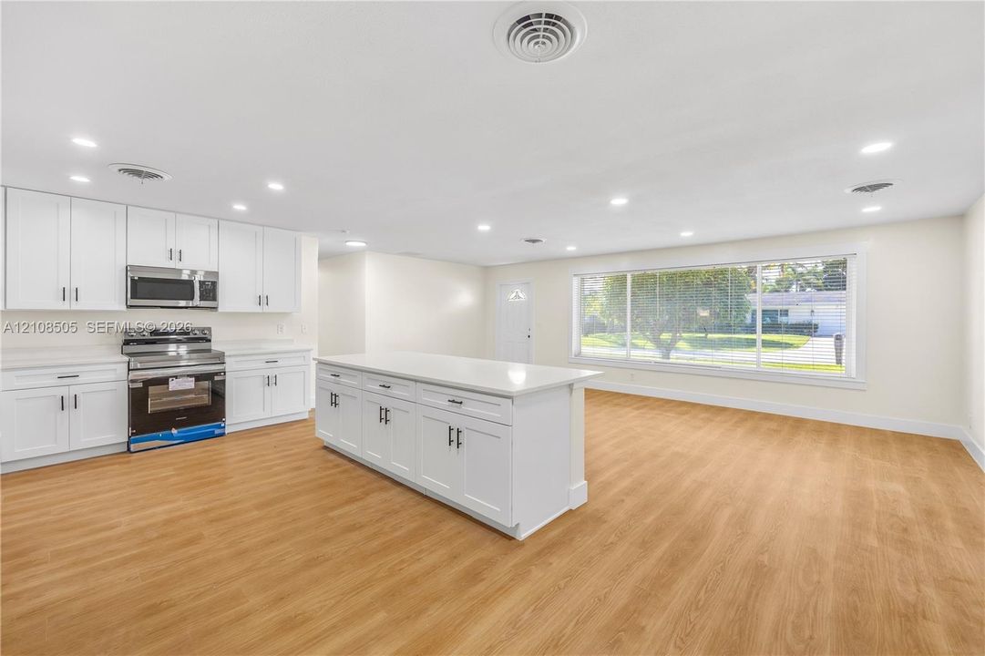Open-concept kitchen featuring white shaker-style cabinetry, a central island with a solid-surface countertop, stainless steel appliances, recessed lighting, and wood-finish flooring