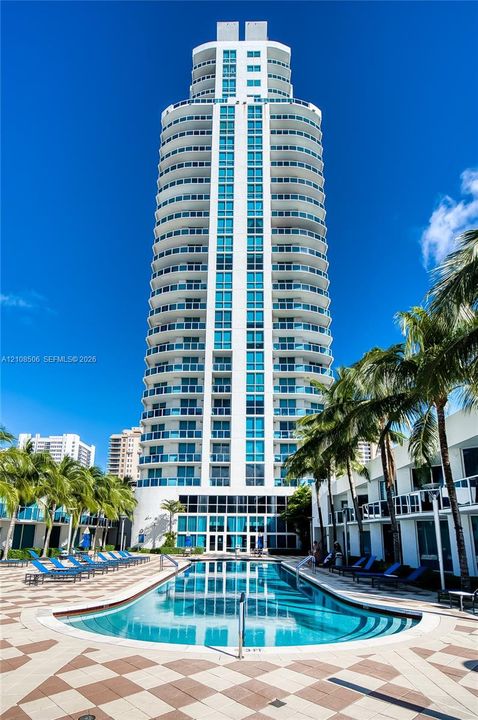 High-rise residential tower featuring wrap-around balconies and floor-to-ceiling glass windows, accented by a centered ground-level pool with tile decking and surrounding palm trees