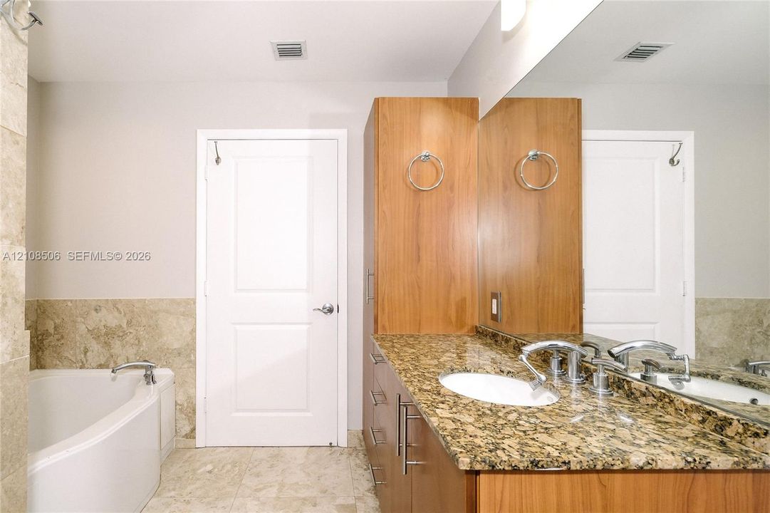 Bathroom featuring stone-surface vanity with dual integrated sinks, floor-to-ceiling wood cabinetry, large wall-mounted mirror, and a soaking tub with stone tile surround