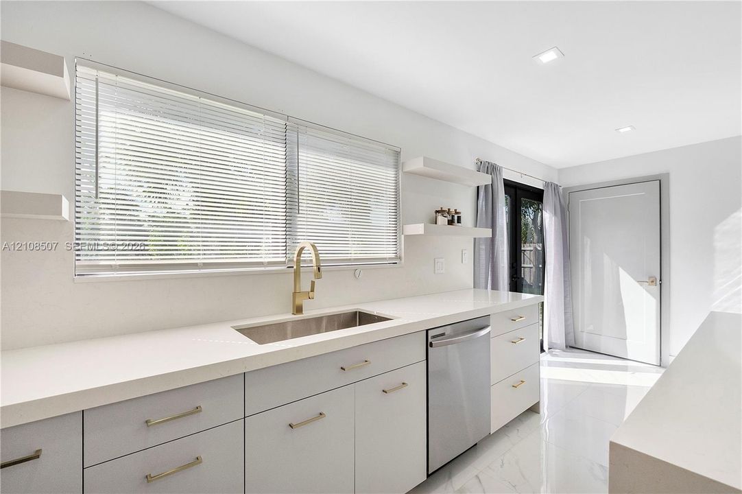 Kitchen featuring light-toned cabinetry, solid surface countertops, a stainless steel undermount sink with a brass-finish faucet, built-in stainless steel dishwasher, and wall-mounted shelving