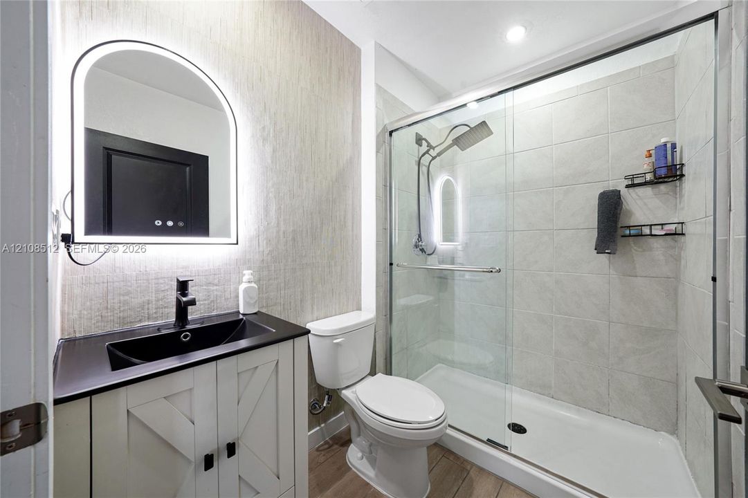 Bathroom featuring a vanity with a dark countertop and integrated sink, arch-top lighted mirror, wood-finish flooring, and a walk-in shower with a glass enclosure and tiled walls